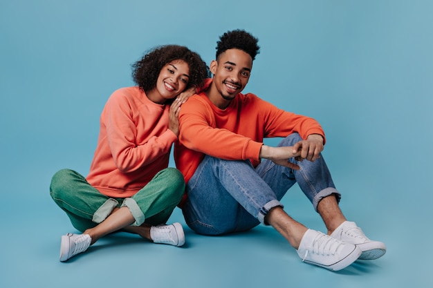 Young man and woman smiling and sitting on blue wall