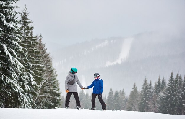 雪に覆われた高い斜面に立って、お互いを見つめている若い男性と女性のスノーボーダー。背景に降雪。