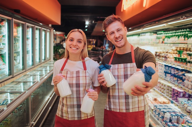 Young man and woman stand in grocery store and dairy shelf. They hold glass bottles of milk and pose on camera. Positive happy workers smiling.