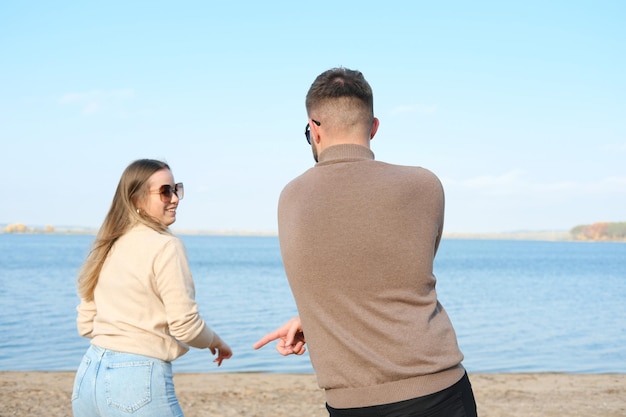 Young man and woman in sunglasses sweater and jeans are having\
fun and dancing on the beach