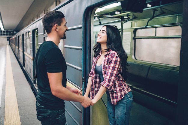 Молодой мужчина и женщина используют под землей. Пара в метро. Молодая брюнетка стоит в подземном вагоне и улыбаются. Она держала мужские руки. Парень стоит на платформе. Люблю стри. Веселый.