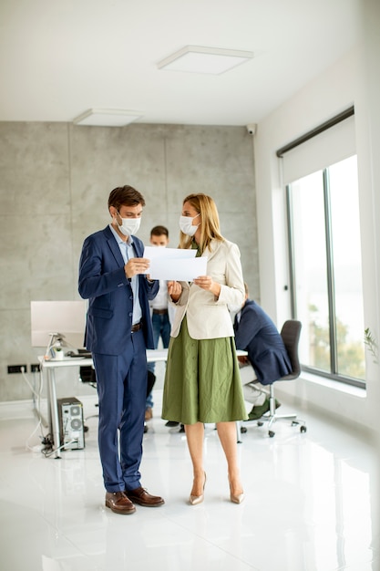 Young man and woman with protective facial masks discussing with paper in hands indoors in the office with young people works behind them