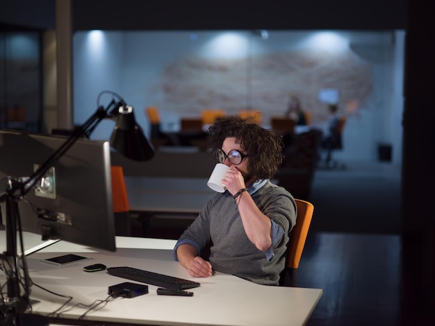 Молодой человек работает за компьютером ночью в темном офисе стартапа. Дизайнер работает в более позднее время.