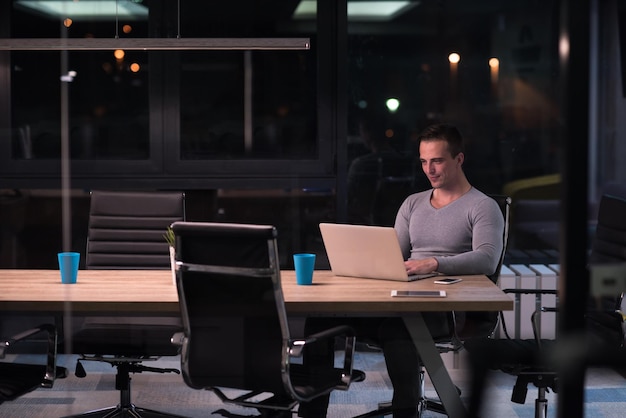 Молодой человек работает на ноутбуке ночью в темном офисе. Дизайнер работает в более позднее время.