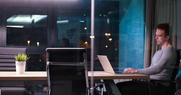 Молодой человек работает на ноутбуке ночью в темном офисе. Дизайнер работает в более позднее время.