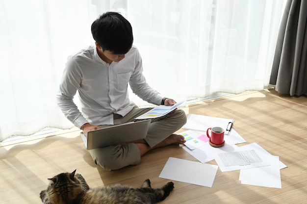 Young man working on a laptop and sitting on the floor in living room.