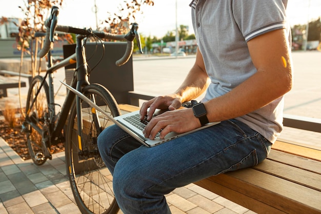 彼の自転車の近くのベンチに座っている間ラップトップに取り組んでいる若い男