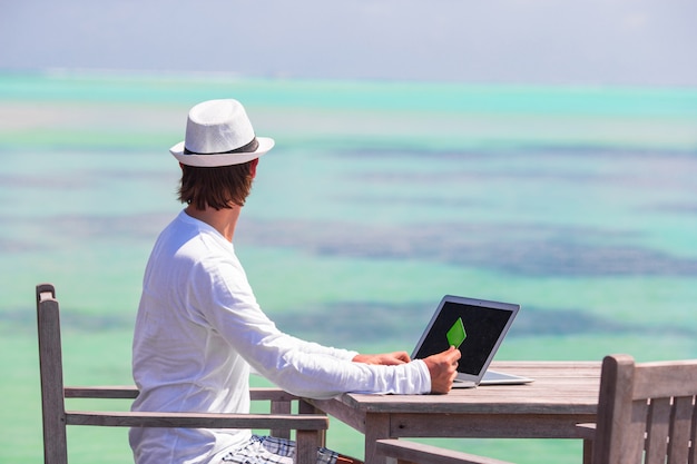 Young man working on laptop with credit card at tropical beach