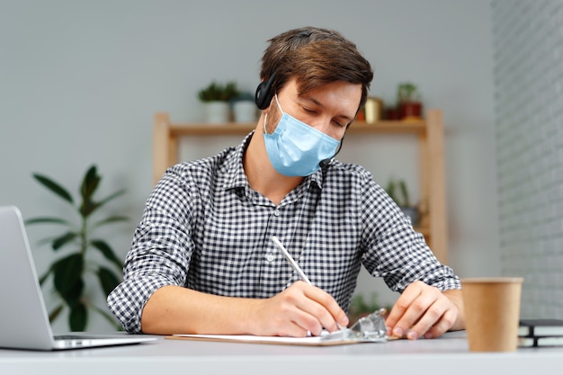 Young man working on laptop with headset and face mask at home