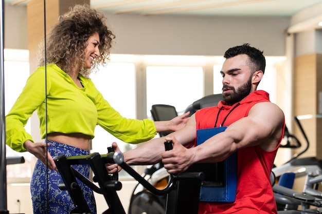 Young Man Working Out In Gym Doing Back Exercise On Machine With Help Of Her Trainer