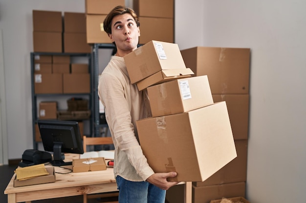 Young man working at small business ecommerce holding packages making fish face with mouth and squinting eyes, crazy and comical.