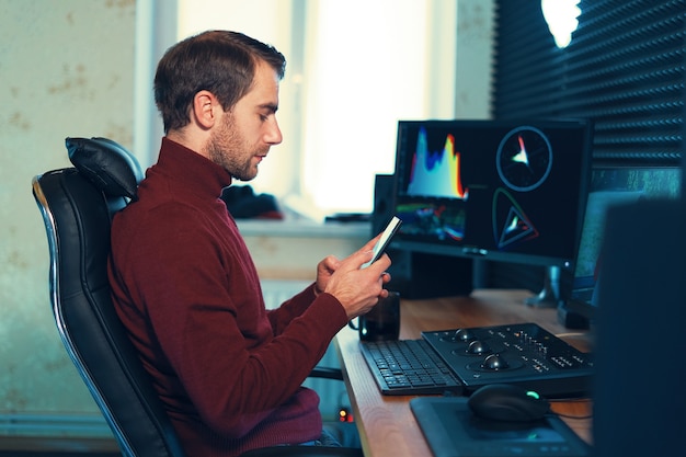 Young man working in the studio using a smartphone and computer. Caucasian freelancer holding mobile phone working on footage, video, design.