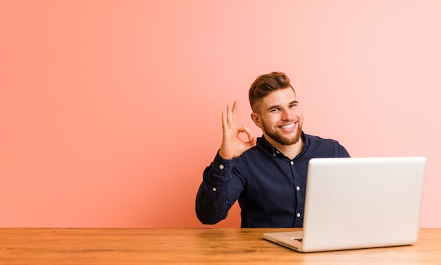 Young man working with his laptop cheerful and confident showing ok gesture.