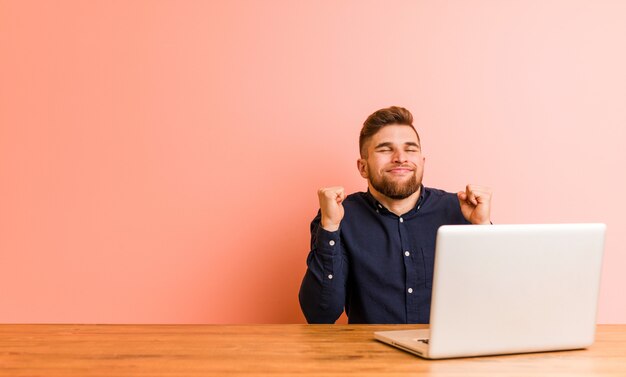 Young man working with his laptop raising fist, feeling happy and successful. Victory concept.