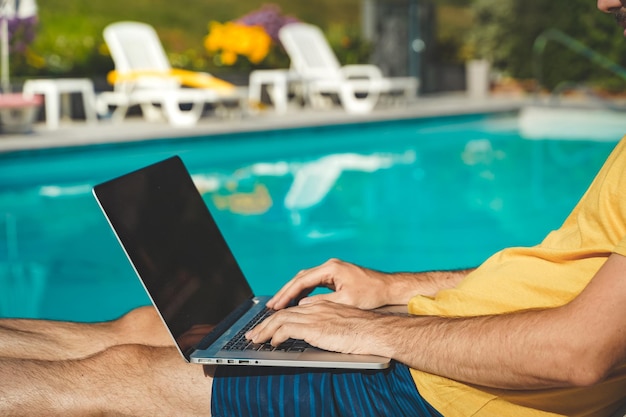 Giovane che lavora con il computer portatile accanto alla piscina durante una giornata di sole uomo attraente che lavora libero professionista