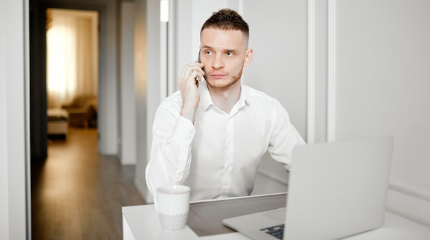 A young man works in the kitchen talks on the phone working at a laptop