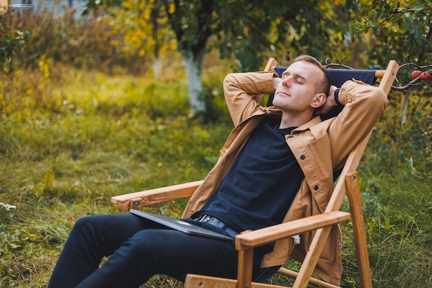 A young man works on a laptop outdoors a young freelancer rests\
in the forest remote work active recreation in the summer tourism\
people concept man sitting on a chair outdoors