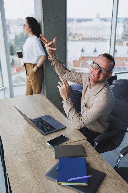 a young man works in the office on a laptop while sitting at the table against the background of colleagues modern workspace with office workers