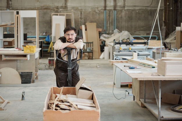 Young man in workwear and eyeglasses leaning on handle of cart with leftovers while carrying them to processing workshop