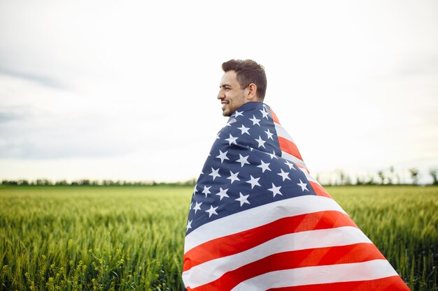 Giovane uomo avvolto nella bandiera americana presso il campo di grano verde