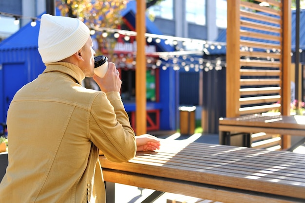 A young man in a yellow jacket sits with his back to the camera drinks coffee and enjoys the good weather