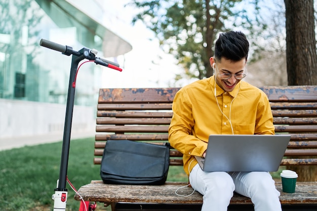 Foto giovane uomo in camicia gialla, studiando in un parco con laptop e scooter