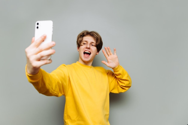 young man in a yellow sweatshirt makes a video call on a smartphone on a background of a gray wall