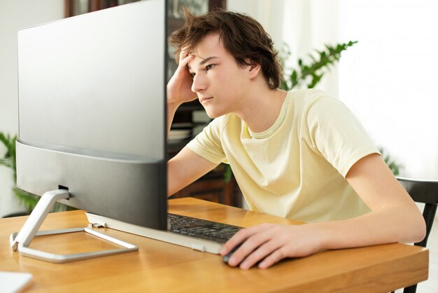 young man in a yellow t-shirt works at home at the computer. chat online. remote work via the Internet on isolation during quarantine of the coronavirus. freelancer