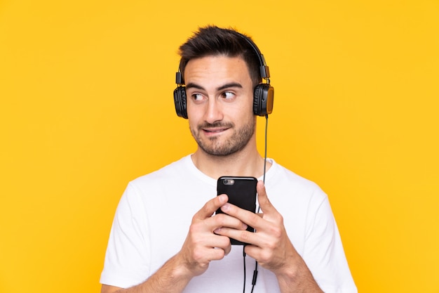 Young man over yellow wall listening music with a mobile and thinking