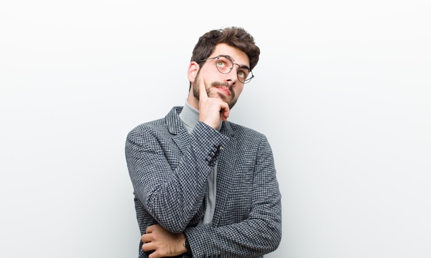 Young manager man with a concentrated look, wondering with a doubtful expression, looking up and to the side against white wall