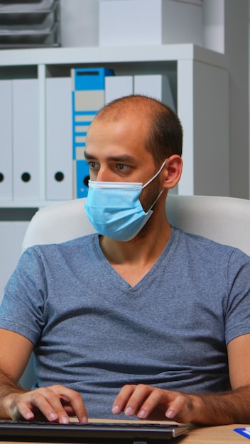 Young manager with protection face mask working alone in office during social distancing. Entrepreneur in new normal personal workplace corporate writing on computer keyboard looking at desktop