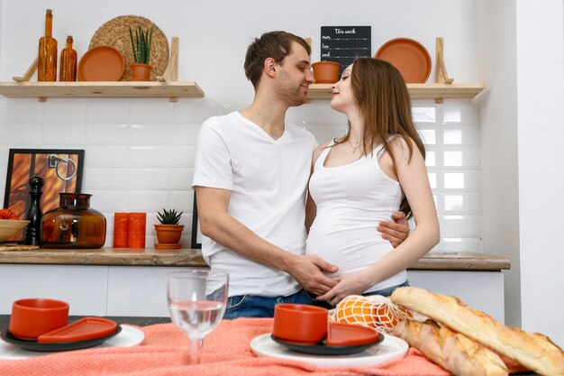 La giovane coppia sposata abbraccia in piedi vicino al tavolo in cucina. il marito abbraccia la moglie incinta, mettendo le mani sulla sua grande pancia. una coppia di innamorati, futuri genitori. stile di vita, persone felici.