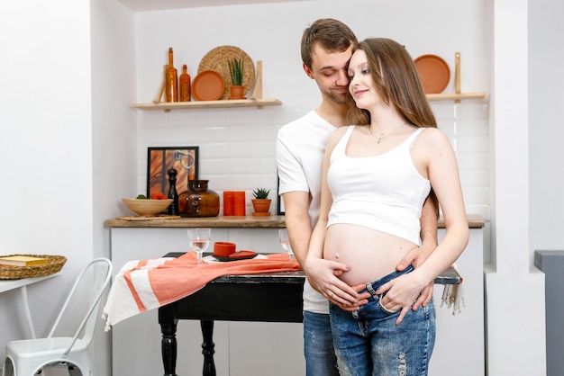 Young married couple embraces standing near table in kitchen.
husband hugs his pregnant wife, putting his hands on her big belly.
a loving couple, future parents. lifestyle, happy people.