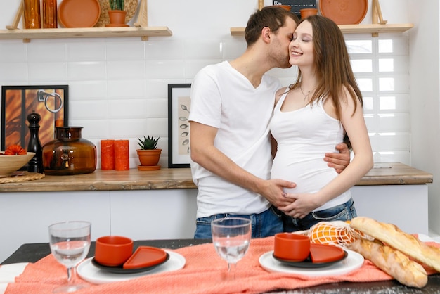 Young married couple embraces standing near table in kitchen\
husband hugs his pregnant wife putting his hands on her big belly a\
loving couple future parents lifestyle happy people