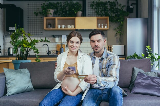 Young married couple husband and pregnant woman sitting together on the couch and hugging using the phone for online shopping in the online store holding a bank credit card