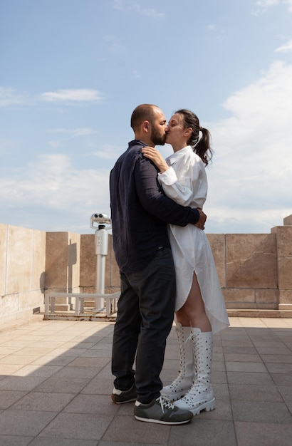 Young married couple kissing during relationship anniversary standing on building rooftop having panoramic view of metropolitan city. View of city skyline from tower observation point.