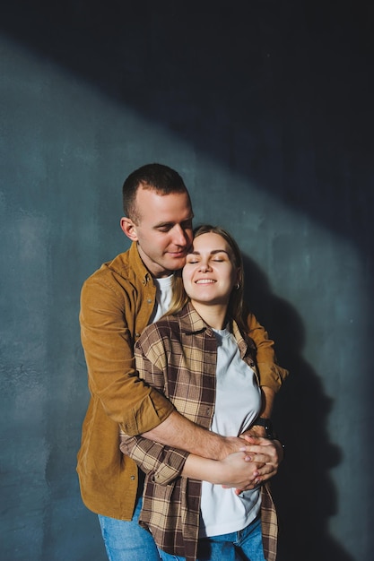A young married couple in love in shirts and jeans on the background of a gray wall The concept of happy family relationships A man and a woman are hugging