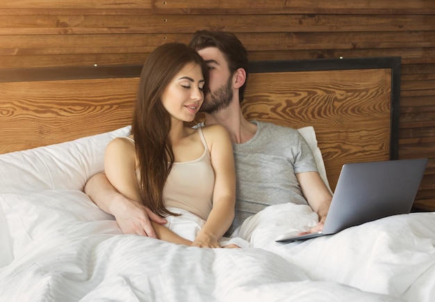 Young married couple lying in bed at home and using laptop.