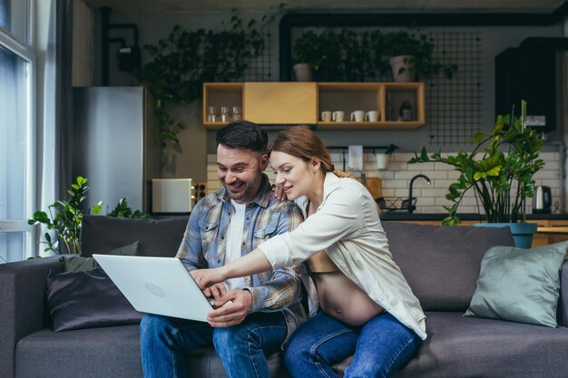 Young married couple man and pregnant woman at home on sofa using laptop for online shopping and video communication