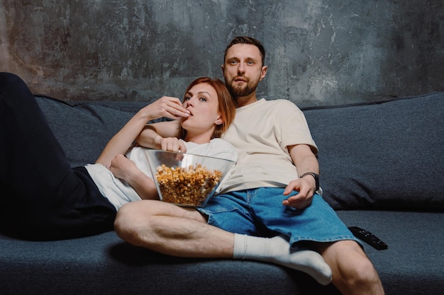 Молодая супружеская пара смотрит телевизор и ест попкорн