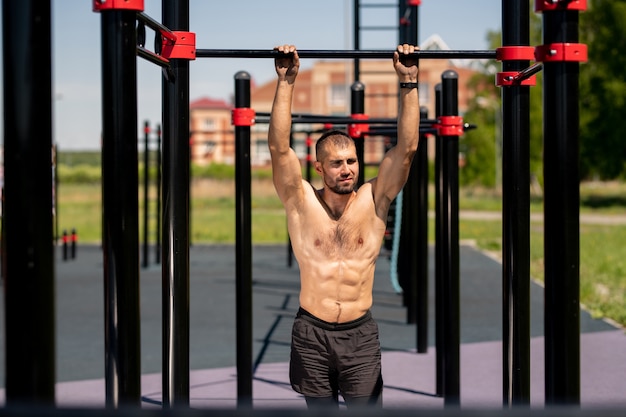 Photo young masculine athlete with mascular torso holding by bars of sports facilities while standing on sportsground