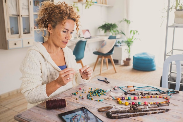 Young mature youthful woman enjoying at home with indoor leisure hobby activity making jewels and necklaces with colorful beads and watching a tablet device to follow tutorial instructions online