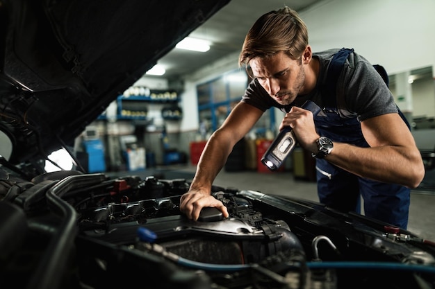 Young mechanic using lamp while examining malfunction of car engine in auto repair shop