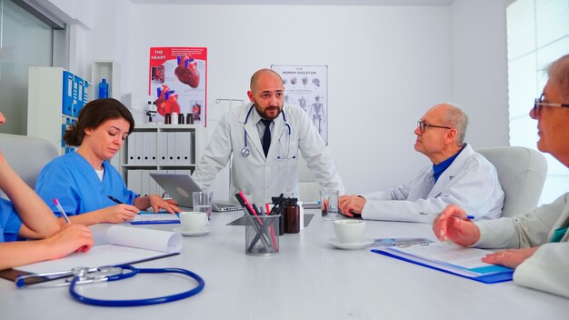 Young medical specialist discussing about patient diagnosis with coworkers in hospital conference room. Clinic expert therapist talking with colleagues about disease, medicine professional