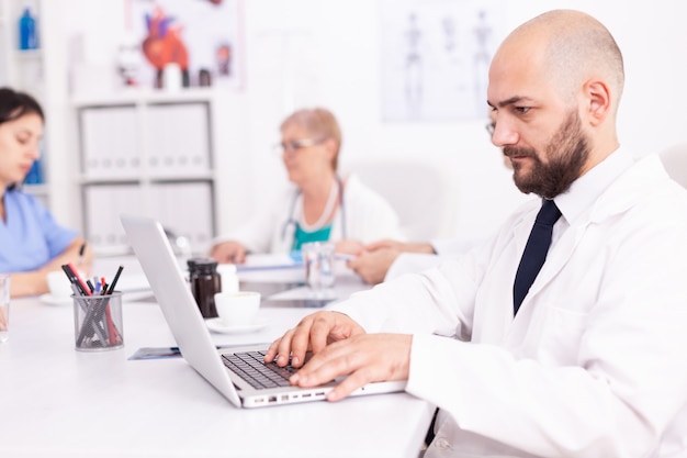 Young medical specialist using laptop in hospital conference room with coworkers. Clinic expert therapist talking with colleagues about disease, medicine professional