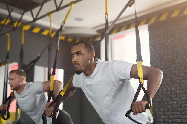 Young men performing TRX training in gym. Muscular guys doing exercise with elastic rope, copy space