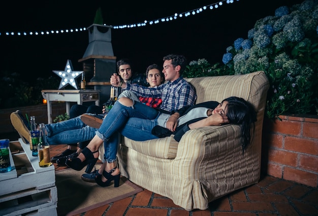 Young men talking and having fun in a outdoors party while woman sleeping lying on sofa. Friendship and celebrations concept.