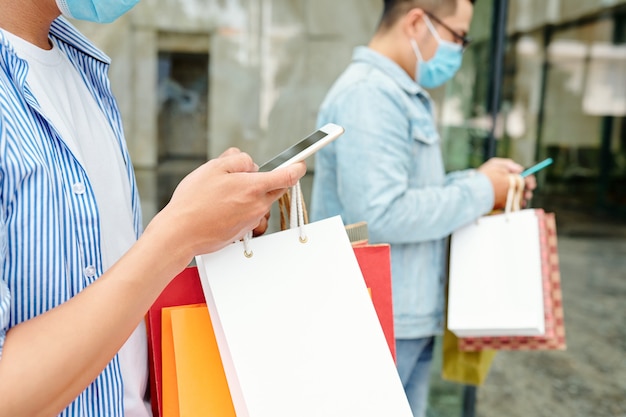 Young men walking outdoors with shopping bags in hands and checking text messages in smartphones