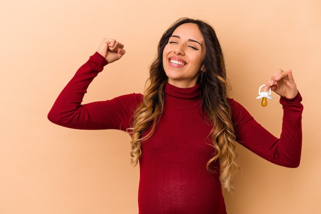 Young mexican pregnant woman holding pacifier isolated on beige background raising fist after a victory, winner concept.