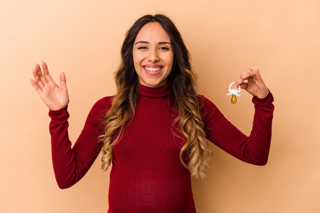 Giovane donna incinta messicana che tiene ciuccio isolato sulla parete beige che riceve una piacevole sorpresa, eccitata e alzando le mani.